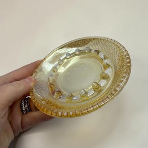 May include: An oval-shaped, iridescent amber glass ashtray. The ashtray features a textured, dotted rim and a central depression with raised, rounded rests for cigarettes. The glass has a subtle, shimmering quality, reflecting light.