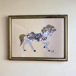 Può includere: Opera d'arte incorniciata raffigurante un cavallo bianco da giostra con sella e dettagli patriottici rossi, bianchi e blu. Il cavallo ha una criniera e una coda fluenti, su sfondo crema. La cornice è dorata.