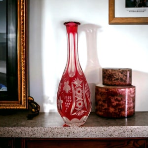 May include: A tall, ruby red glass vase with intricate etched floral and starburst designs. The vase has a slender neck and a wide, rounded body. The vase is displayed on a countertop with two decorative containers.