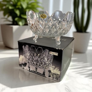 May include: A clear, diamond-cut crystal bowl with three small feet, sitting atop a black box. The bowl has a scalloped rim and a small gold sticker. The box features an image of the bowl and the word "Triumph".