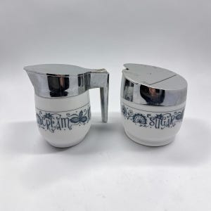 May include: A vintage cream and sugar set. The set includes a cream pitcher with a handle and a sugar bowl, both with chrome lids. The white ceramic pieces have blue floral and scroll designs, with the words "CREAM" and "SUGAR" in blue.