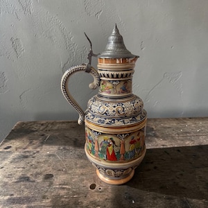 May include: A ceramic beer stein featuring a pointed metal lid with a hinge and a curved handle. The body is decorated with colorful painted scenes of figures in historical attire, intricate scrollwork, and relief patterns in blue, gold, and tan.