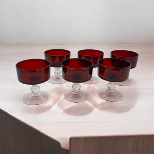 May include: Set of six red glass dessert dishes with clear glass stems and bases. The dishes are arranged in a row on a light wood surface.