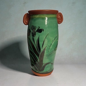 May include: A handcrafted ceramic vase with a green glaze and a brown rim and base. The vase features a black floral design and two brown handles. The vase is approximately 25 cm tall.