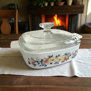 May include: A white casserole dish with a clear glass lid. The dish features a floral pattern with yellow, pink, and blue flowers and fruits. The dish is on a white cloth on a wooden table.