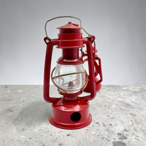 May include: A bright red metal hurricane lantern featuring a clear glass globe protected by a wire cage. The lantern has a top handle for carrying and a circular base with a side opening. A second, smaller red lantern is visible behind it.
