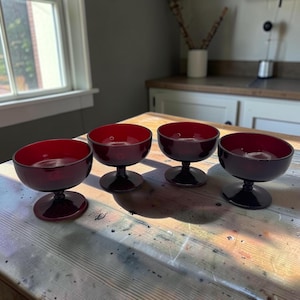 May include: A set of four matching ruby red glass dessert bowls, each featuring a wide, shallow basin, a short stem, and a circular pedestal base. The glass is translucent and deep red in color, with a smooth, polished finish throughout.