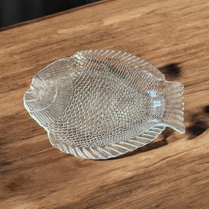Op de afbeelding: Een heldere, glazen schaal in de vorm van een vis, met gedetailleerde schubben- en vinnenpatronen. De schaal is ontworpen om op een vis te lijken en is op een houten oppervlak geplaatst. De schaal wordt waarschijnlijk gebruikt om voedsel te serveren of als decoratief item.