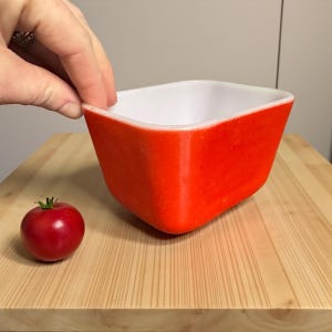 May include: A square, orange and white bowl with a tomato on a wooden cutting board. The bowl has a bright orange exterior and a white interior. The tomato is red and round. The bowl is being held by a hand.