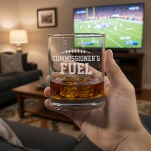Puede incluir: Un vaso transparente lleno de líquido ámbar y cubitos de hielo, sostenido en la mano de una persona. El vaso está grabado con las palabras "COMMISSIONER'S FUEL" en blanco, con un gráfico de fútbol americano encima. Un televisor que muestra un partido de fútbol americano es visible en el fondo.