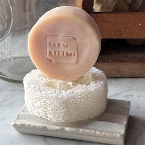 May include: A round, light pink soap bar with the text "FARM KITTEN" stamped on it, resting on a white loofah sponge and a white ceramic soap dish. The soap dish has a rectangular shape with ridges.