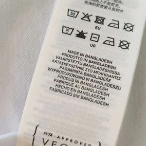 May include: A white fabric tag with care symbols and text in multiple languages, including "MADE IN BANGLADESH" and "PETA-APPROVED VEGAN". The tag also features washing instructions.