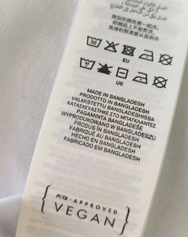 May include: A white fabric tag with care symbols and text in multiple languages, including "MADE IN BANGLADESH" and "PETA-APPROVED VEGAN". The tag also features washing instructions.