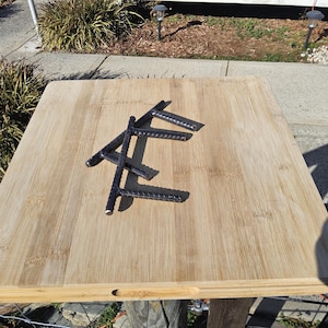 May include: A light-colored wooden cutting board with dark metal rebar pieces arranged on top. The rebar is in a geometric pattern. The cutting board is square and has a natural wood grain.
