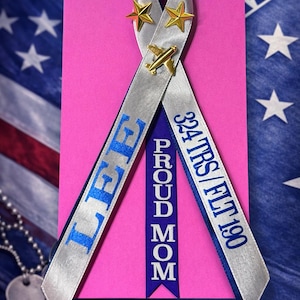 May include: A pink card with a silver ribbon featuring the text "LEE" and "324 TRS/FLT 190" in blue. The ribbon is adorned with gold stars and a small airplane. The words "PROUD MOM" are on a blue ribbon.