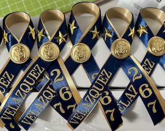 May include: Six navy blue and gold ribbon favors with a gold anchor charm and the text "VELAZQUEZ" and a number. The favors are tied in a bow.