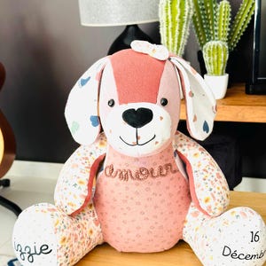 May include: A plush dog toy with a pink and floral pattern. The dog has a black heart-shaped nose, embroidered text "amour", and the name "Izzie" and the date "December 16, 2022" embroidered on its paws. The dog has a small bow on its head.
