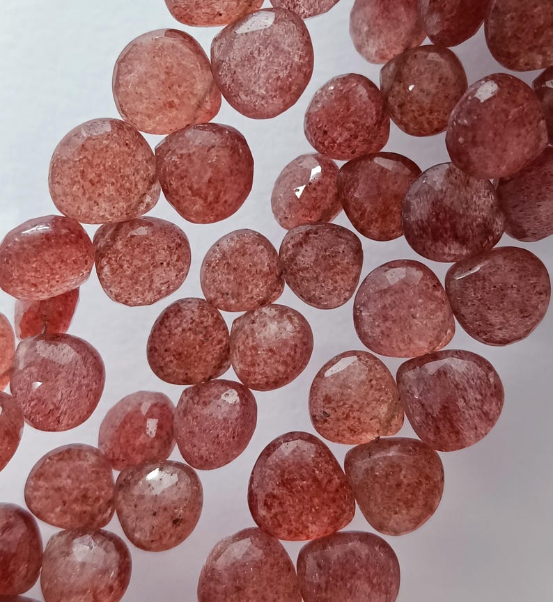 May include: A strand of pink and red gemstone beads. The beads are faceted and have a slightly irregular shape. They are strung on a clear thread.