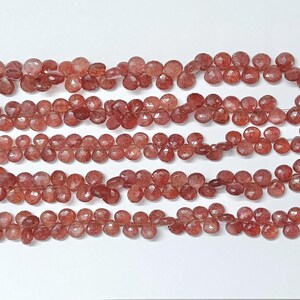 May include: A strand of red gemstone beads with a faceted, heart-shaped design. The beads are strung on a cord with pink tassels at each end.