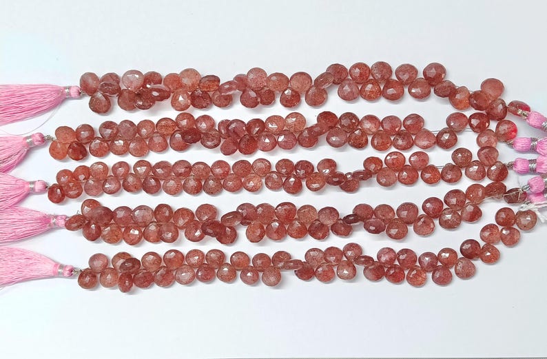 May include: A strand of red gemstone beads with a faceted, heart-shaped design. The beads are strung on a cord with pink tassels at each end.
