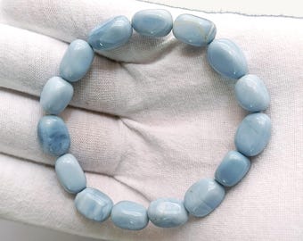 May include: A stretch bracelet composed of smooth, polished, light blue oval-shaped beads. The beads exhibit a consistent pale blue color with subtle variations in tone and a glossy finish, arranged in a circular pattern on a white fabric surface.
