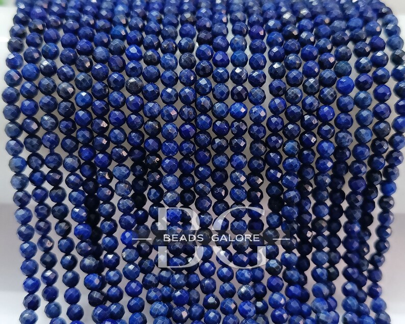 May include: A strand of deep blue faceted lapis lazuli beads. The beads are small and round, and they have a smooth, polished finish. "BEADS GALORE" is printed on a white banner.