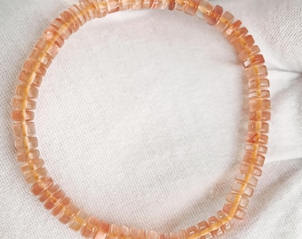 May include: A circular bracelet composed of small, translucent, peach-colored disc-shaped beads. The beads are strung together on a thin, visible orange cord, creating a uniform, repeating pattern of light orange and pinkish-toned circular segments.