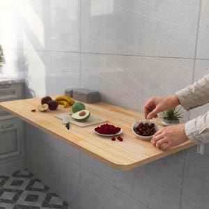 May include: A light-colored, wall-mounted wooden folding table with rounded corners. The table is set with various fruits, including apples, bananas, and raspberries. A person is arranging cherries in a small white bowl. The table is mounted on a gray tiled wall.