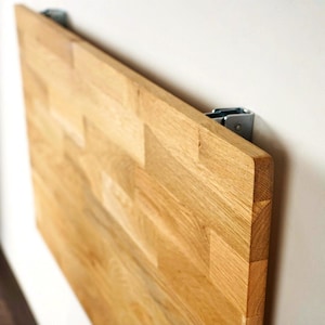 May include: A rectangular, light-colored wooden table, mounted on a white wall with metal hinges. The table is made of multiple wood pieces, creating a textured surface. The wood grain is visible, and the table is angled.
