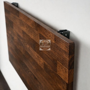 Peut inclure: Une table pliante murale rectangulaire marron foncé. La table est en bois et présente un motif de grain naturel. La table est fixée au mur avec des supports métalliques noirs. Le logo "WoodySnap" est visible sur la table.