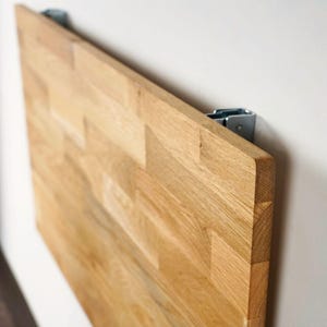 Puede incluir: Una mesa rectangular de madera clara, montada en la pared, con un patrón de vetas de madera natural. La mesa está fijada a una pared blanca con soportes metálicos. La mesa está hecha de varias piezas de madera.