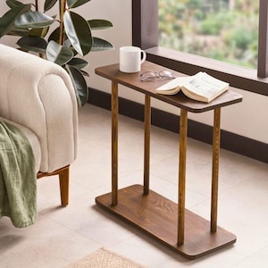 Könnte beinhalten: Ein dunkelbrauner Beistelltisch aus Holz, ca. 100 cm lang, mit rechteckiger Ober- und Unterseite, getragen von drei zylindrischen Beinen. Eine weiße Tasse, eine Brille und ein offenes Buch liegen auf dem Tisch.