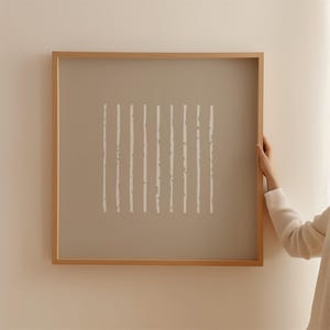 May include: A square wall art piece featuring a light brown background with a series of eleven vertical white lines. The lines have a textured, irregular appearance and are enclosed within a simple, light-toned wooden frame.