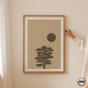 K&ouml;nnte beinhalten: Ein gerahmter Kunstdruck mit der dunkelbraunen Silhouette eines Baumes und einem massiven dunkelbraunen Kreis, der die Sonne darstellt, vor einem strukturierten beigen Hintergrund. Der Druck ist in einem hellen Holzrahmen mit wei&szlig;em Passepartout.