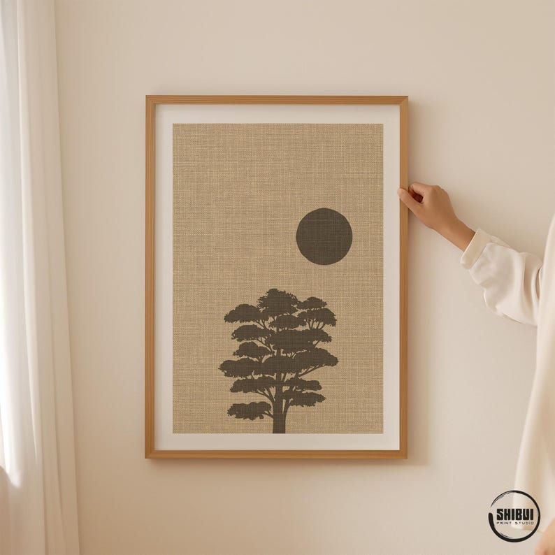 K&ouml;nnte beinhalten: Ein gerahmter Kunstdruck mit der dunkelbraunen Silhouette eines Baumes und einem massiven dunkelbraunen Kreis, der die Sonne darstellt, vor einem strukturierten beigen Hintergrund. Der Druck ist in einem hellen Holzrahmen mit wei&szlig;em Passepartout.