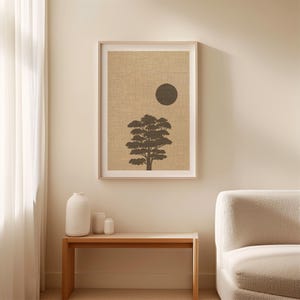 K&ouml;nnte beinhalten: Ein gerahmter Wandkunstdruck mit der dunkelbraunen Silhouette eines Baumes, zentriert unter einem massiven dunkelbraunen Kreis auf einem strukturierten beigen Hintergrund. Das Kunstwerk ist in einem hellen Rahmen und h&auml;ngt an einer Wand &uuml;ber einem Holztisch.