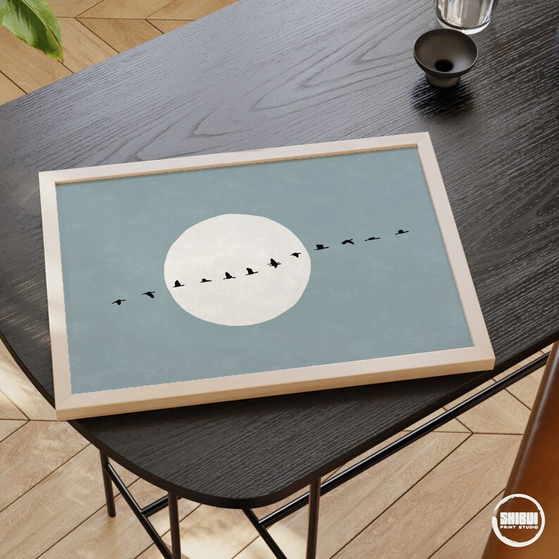 May include: A rectangular framed wall art print featuring a light blue background with a large, solid white circle representing a moon. A horizontal line of small black bird silhouettes flies across the center of the moon and the blue sky.