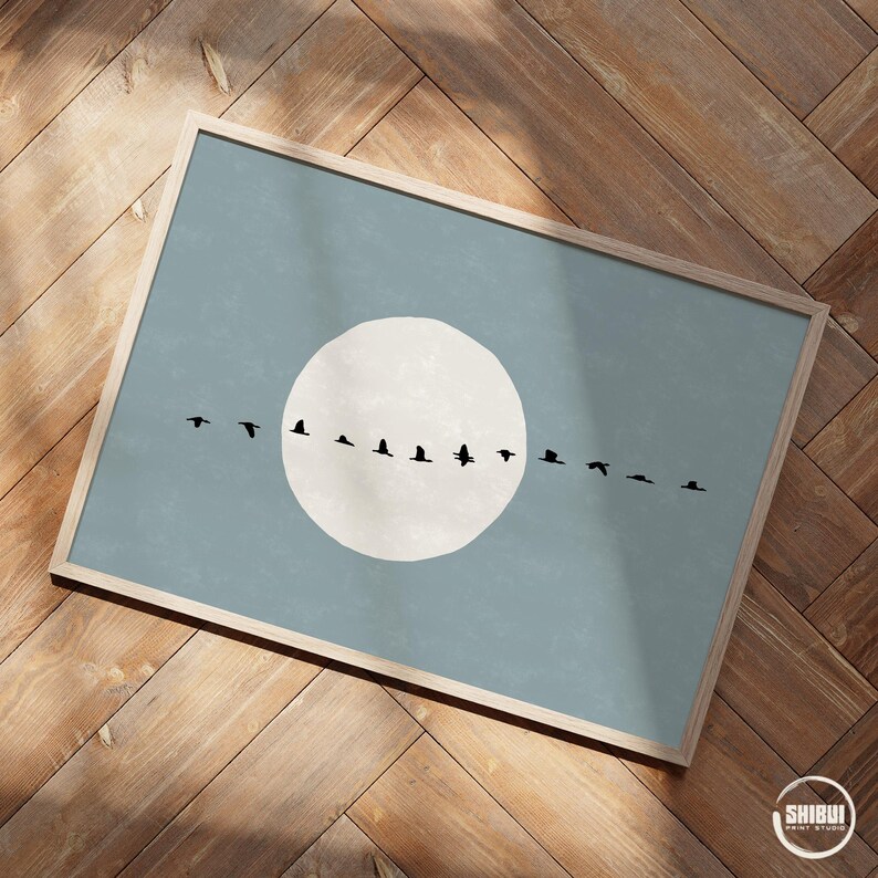 May include: A rectangular framed wall art print featuring a large, solid off-white circle centered on a muted blue background. A horizontal line of twelve black bird silhouettes in various flight positions crosses the center of the circle.