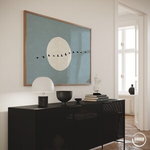May include: A rectangular wall art print in a wooden frame featuring a large, pale beige circle centered on a muted blue background, with a horizontal line of small black bird silhouettes flying across the middle of the circle.