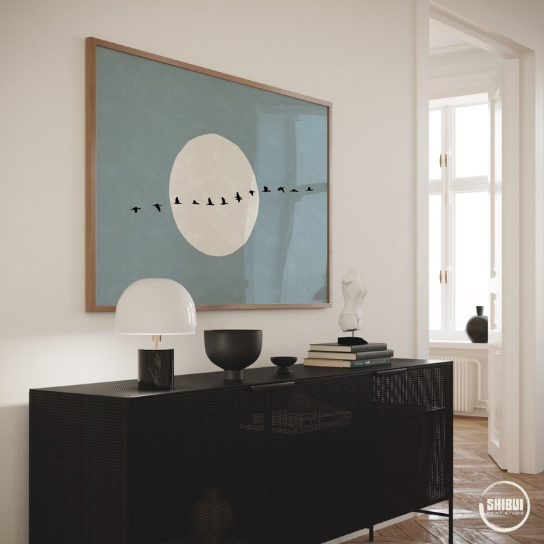 May include: A rectangular wall art print in a wooden frame featuring a large, pale beige circle centered on a muted blue background, with a horizontal line of small black bird silhouettes flying across the middle of the circle.
