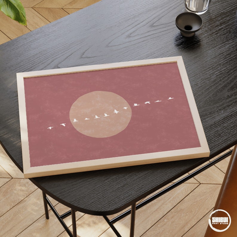 May include: A framed print with a minimalist design featuring a large, peach-colored circle and a line of white birds flying across the center. The background is a muted pink.