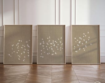 May include: A set of three rectangular wall art prints featuring a beige, textured linen-look background with a scattered pattern of small, white, flying bird silhouettes. Each print is encased in a thin, light-colored wooden frame.