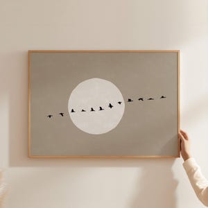 May include: A rectangular wall art print in a light wooden frame featuring a taupe background with a large, pale off-white circular moon shape in the center. A horizontal line of small, black bird silhouettes flies across the moon and background.