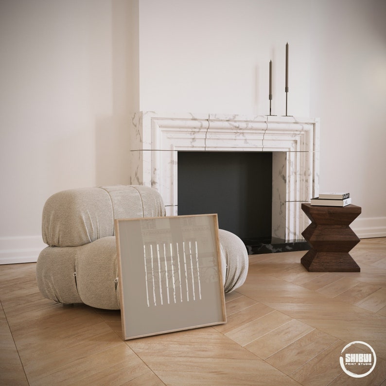 May include: A square framed print featuring a taupe background with a series of vertical white dashed lines. The artwork is positioned on a light wood floor in front of a beige upholstered armchair, a marble fireplace, and a dark wooden side table.