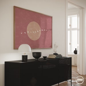 May include: A framed abstract print with a pink background, a large beige circle, and a line of white birds flying across the center. The print is hanging on a white wall above a black dresser with a lamp, bowl, and books.