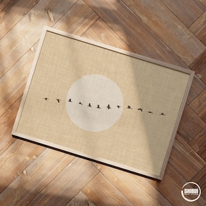 May include: A framed print featuring a beige background with a white circle and a line of black bird silhouettes. The print is in a light-colored wooden frame, resting on a wooden surface. The artwork evokes a sense of nature and flight.