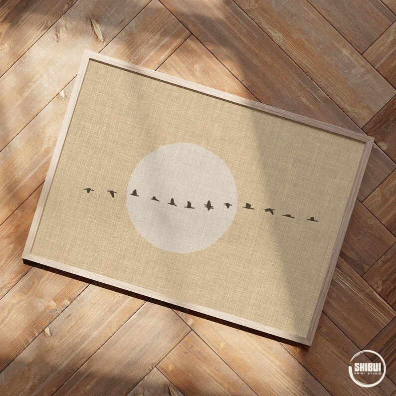 May include: A framed print featuring a beige background with a white circle and a line of black bird silhouettes. The print is in a light-colored wooden frame, resting on a wooden surface. The artwork evokes a sense of nature and flight.
