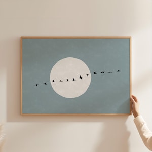 May include: A rectangular wall art print in a light wood frame featuring a muted blue background with a large, off-white circular moon shape in the center. A horizontal line of small, black bird silhouettes flies across the moon and sky.