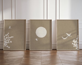 May include: A set of three rectangular framed wall art prints featuring a textured beige background. The prints display white silhouettes of flying cranes, a large circular moon, and flowering tree branches arranged in a triptych composition.