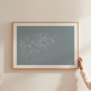 Op de afbeelding: Een ingelijst kunstwerk aan de muur met een getextureerde, leiblauwe achtergrond en een verspreid patroon van kleine, witte, gestileerde vogels in vlucht. Het kunstwerk zit in een lichtgekleurde houten lijst met een witte passe-partout.